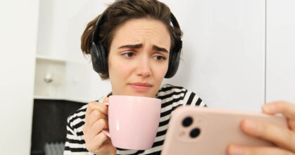 Portrait of woman with sad face, watching something on mobile phone, social media app, drinking tea, wearing wireless headphones